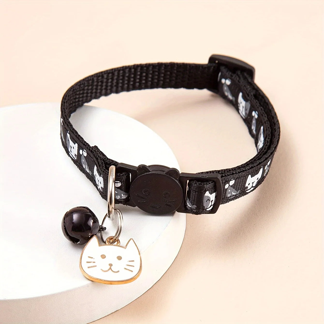 Collier noir pour chat avec une clochette et un charme en forme de visage de chat sur fond beige

