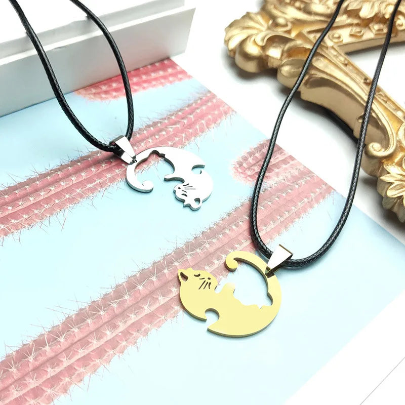 Deux pendentifs en forme de chat sur des cordons noirs avec un cadre décoratif doré en arrière-plan.

