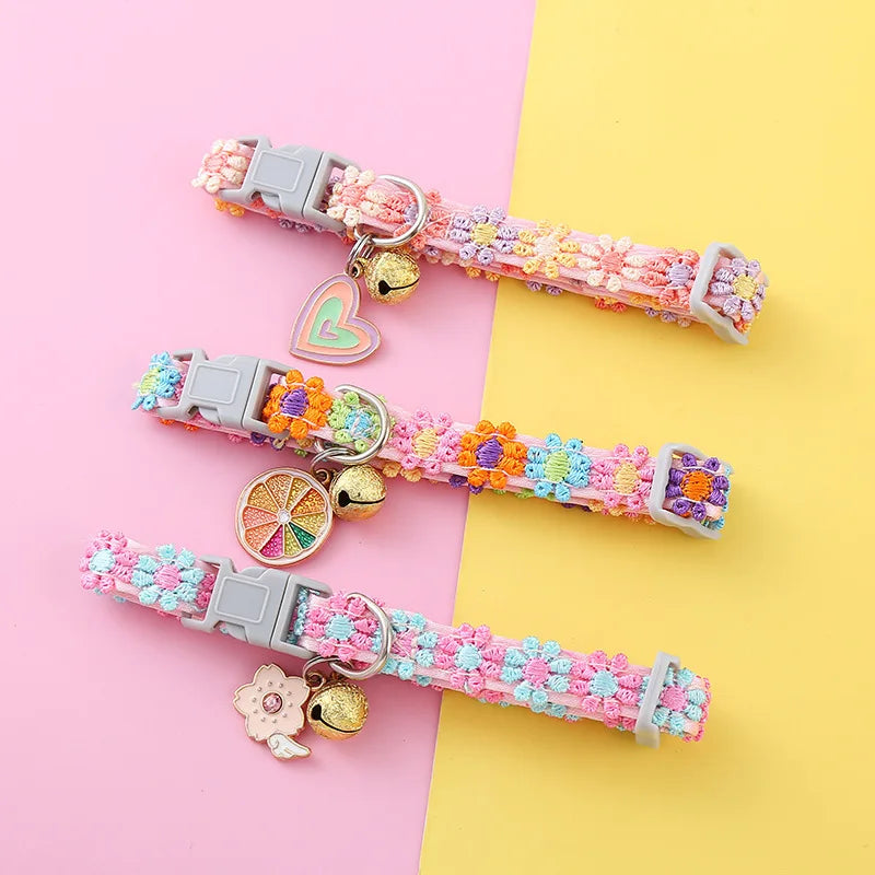 Trois colliers colorés pour animaux avec des motifs floraux et des breloques sur fond rose et jaune

