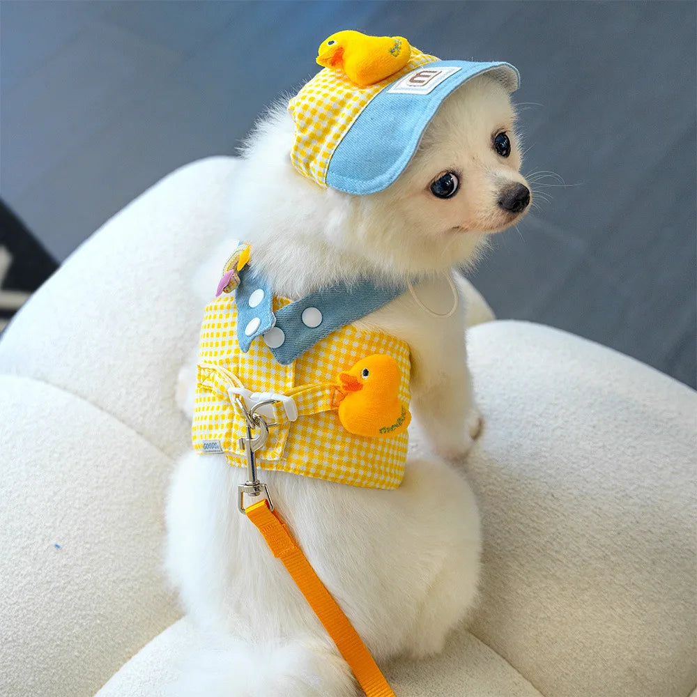 Petit chien blanc portant une tenue jaune à carreaux avec des décorations de canard et une casquette bleue.

