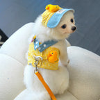 Petit chien blanc portant une tenue jaune à carreaux avec des décorations de canard et une casquette bleue.

